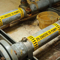 Two larges pipes labeled with bright yellow pipe markers warning of caustic liquid.