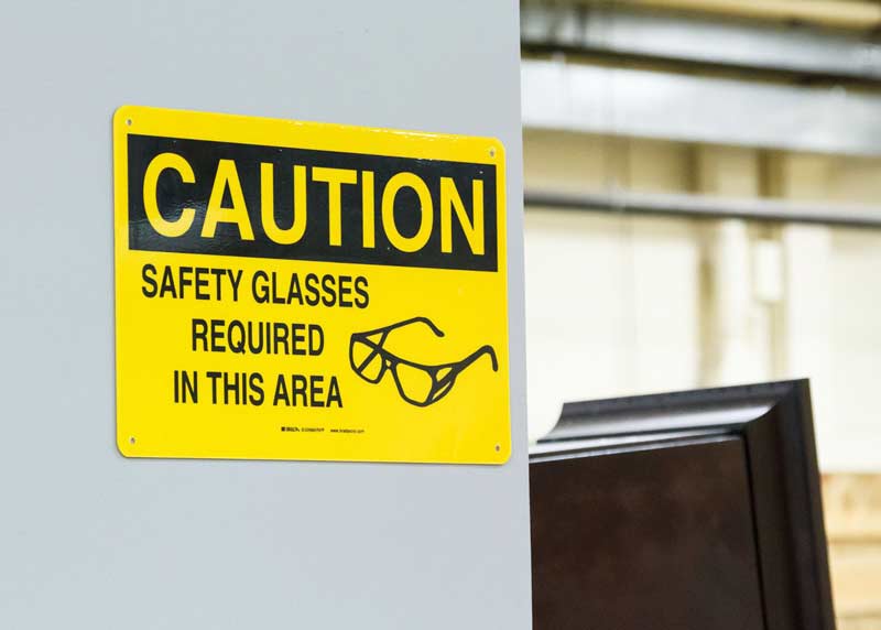 A yellow caution safety sign with an image of safety glasses with text telling readers that safety glasses are required in that area.