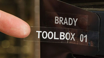 A clear label with white writing that reads Brady toolbox 01 being applied to a piece of metal.
