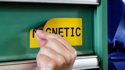 A sample of Brady's magnetic-polyester label material with the text MAGNETIC printed on it magnetically adhered to the metal cover of a piece of equipment.