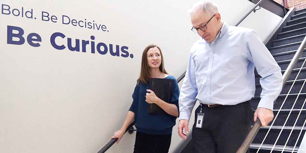 A man and a woman are having a conversation as they walk down a stairwell. The wall next to them has lettering that reads "Be bold. Be decisive. Be curious."